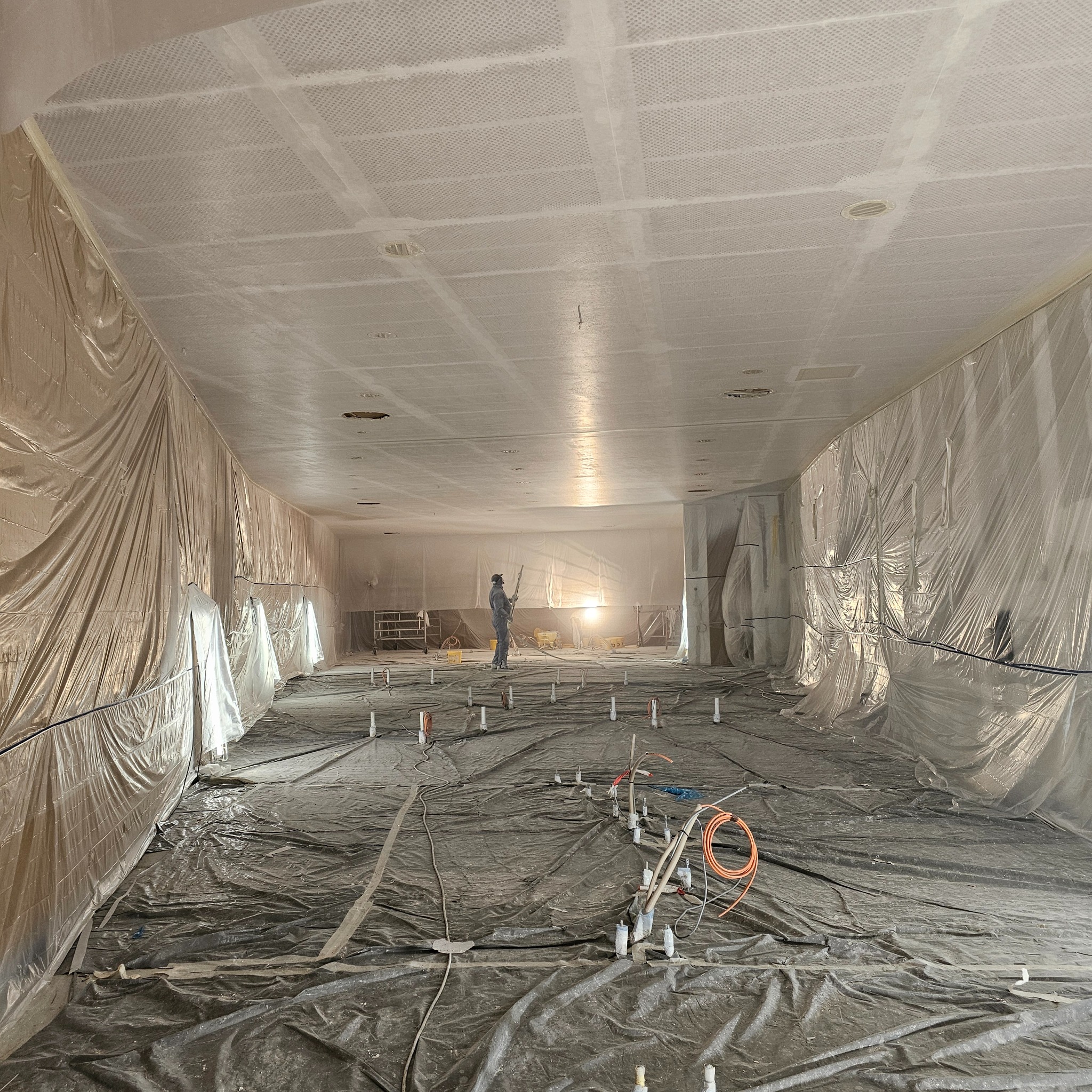 Sufit z perforacją, przygotowany do malowania natryskowego. Ściany i podłoga zabezpieczone folią. Osoba w kombinezonie maluje sufit. Widoczne instalacje podłogowe.