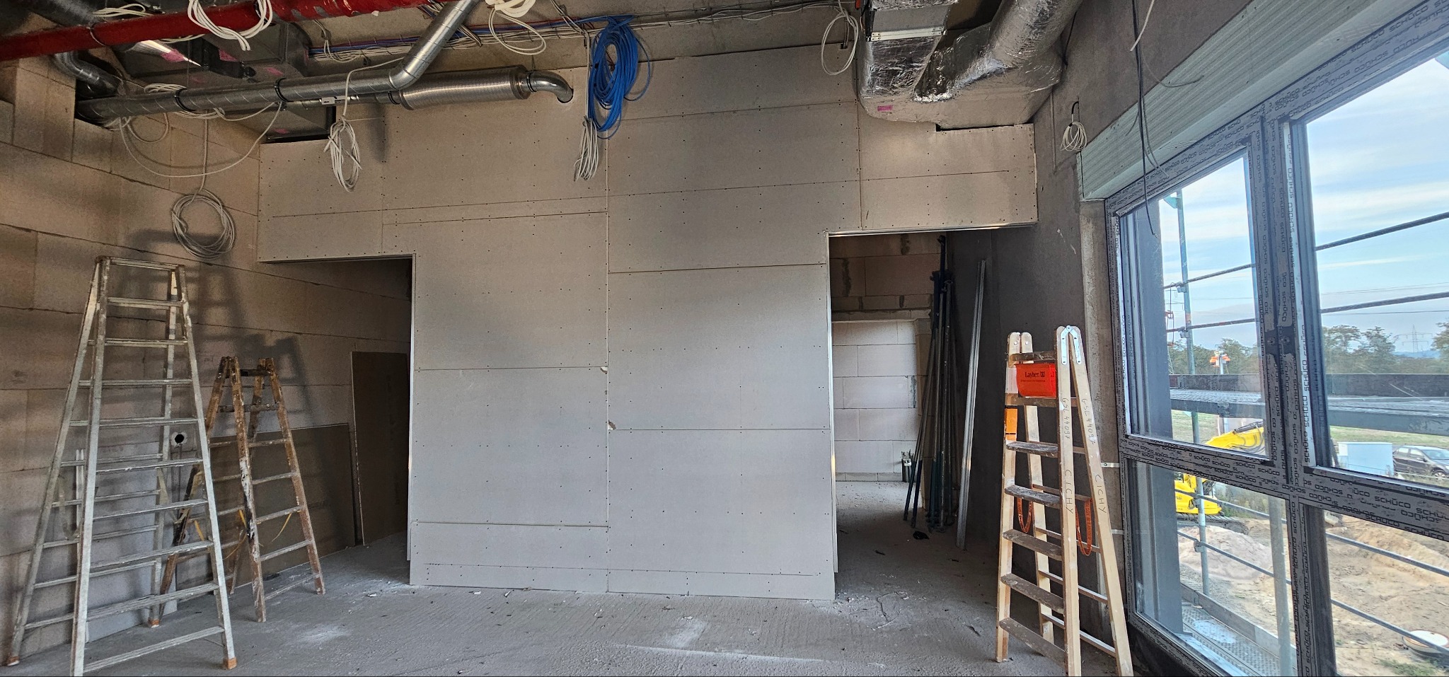 Wnętrze w trakcie prac z płytami karton-gips, widoczne drabiny, instalacje pod sufitem i okno z widokiem na zewnątrz. Ściany częściowo wykończone.