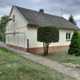 Dwukondygnacyjny dom po odświeżeniu elewacji w jasnych barwach, z widocznymi rusztowaniami i ozdobnym, ciemniejszym pasem u dołu. Na pierwszym planie zadbany trawnik.