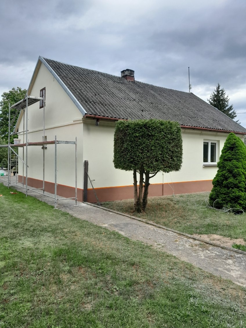 Dwukondygnacyjny dom po odświeżeniu elewacji w jasnych barwach, z widocznymi rusztowaniami i ozdobnym, ciemniejszym pasem u dołu. Na pierwszym planie zadbany trawnik.