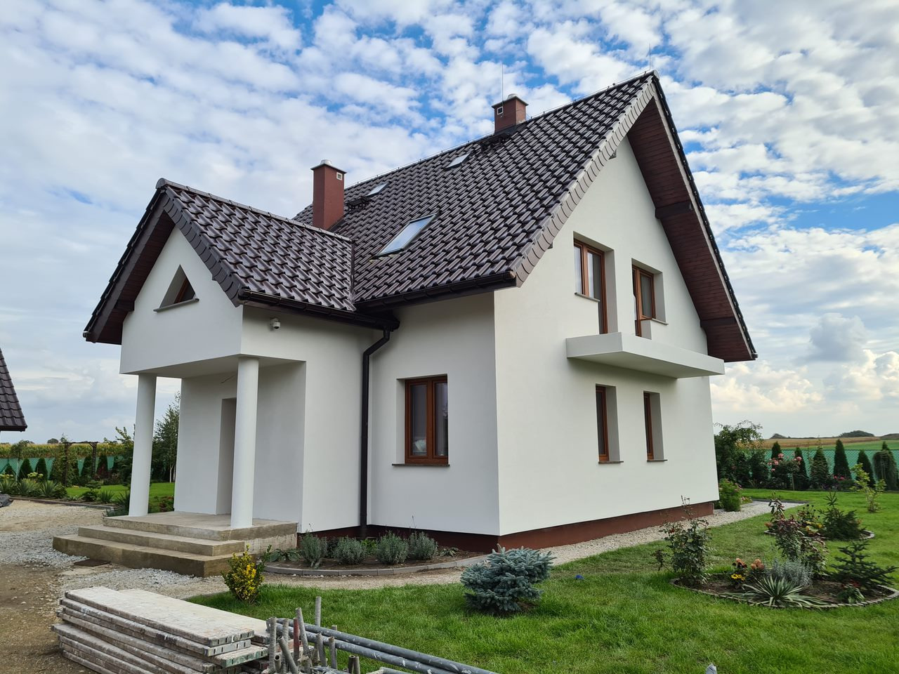 Elewacja domu jednorodzinnego z brązowym dachem i oknami, biała fasada, widok z ogrodu w słoneczny dzień. Na pierwszym planie materiały budowlane.