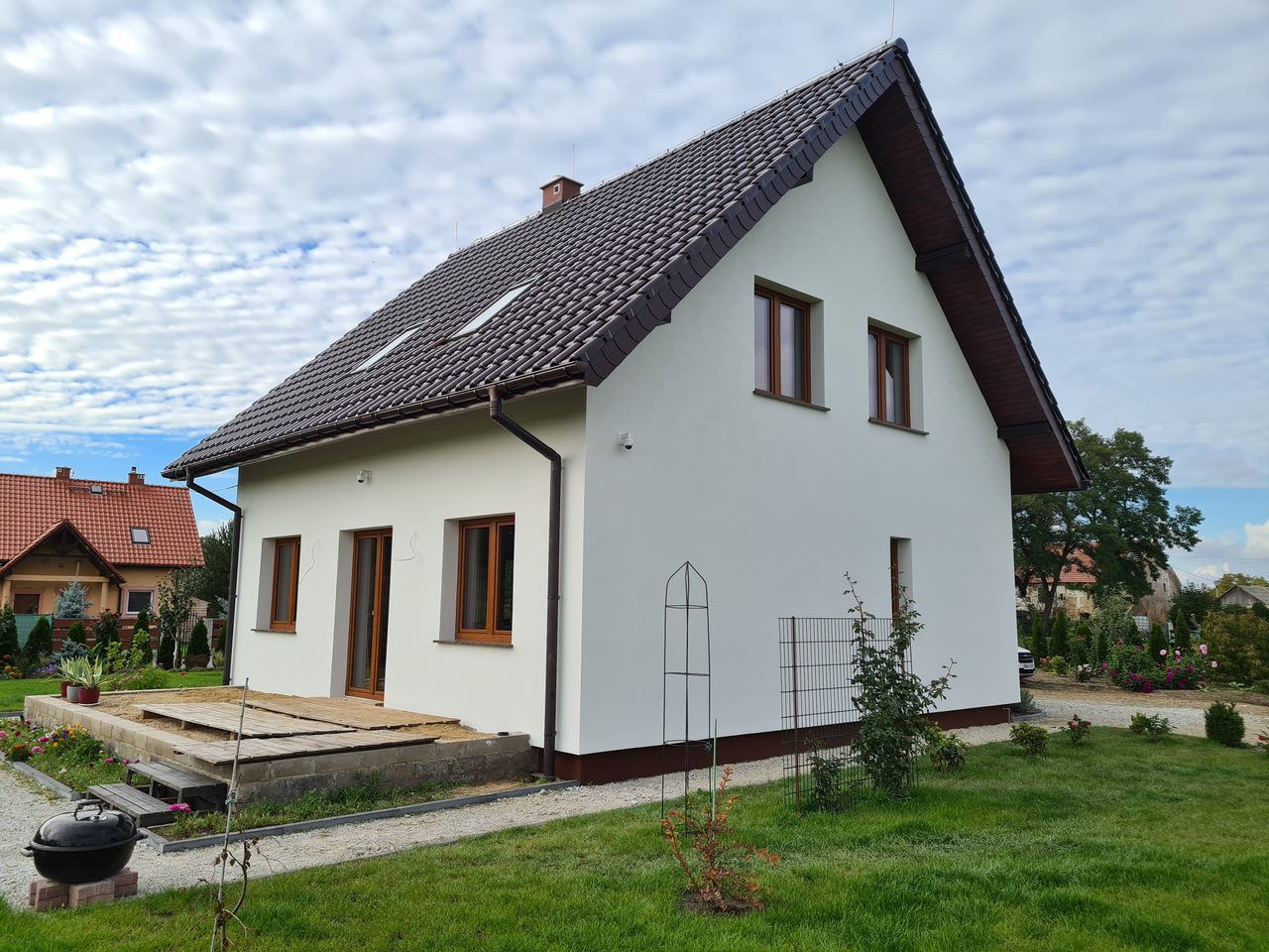 Biały dom jednorodzinny z brązowym dachem, drewnianą podbitką i oknami. Widoczny taras w budowie, trawnik i fragment ogrodu. Ujęcie w słoneczny dzień.