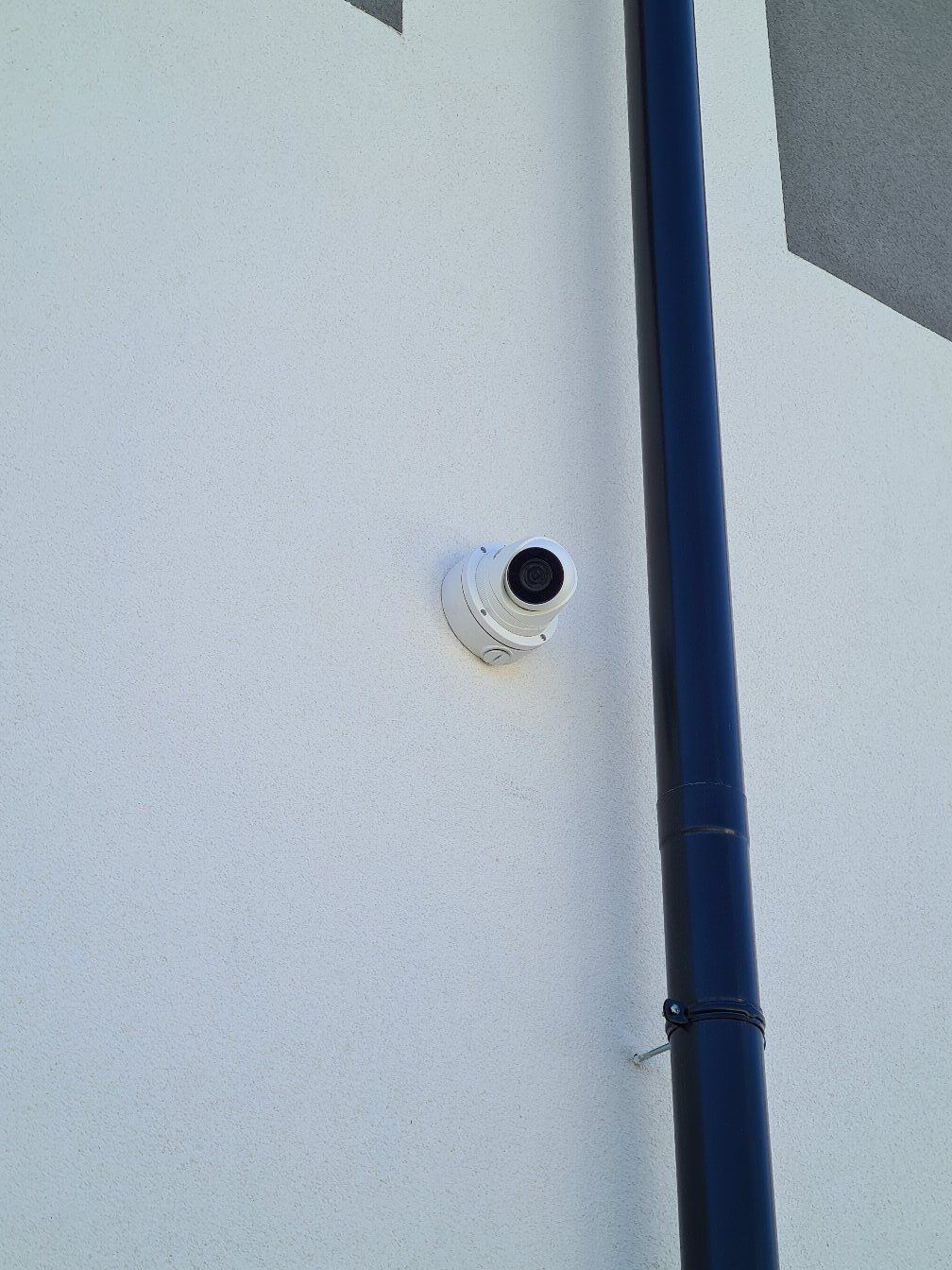 Kamera monitoringu kopułkowa zamontowana na jasnej elewacji budynku, obok rynny spustowej. Widok z dołu, podkreślający dyskretny montaż.