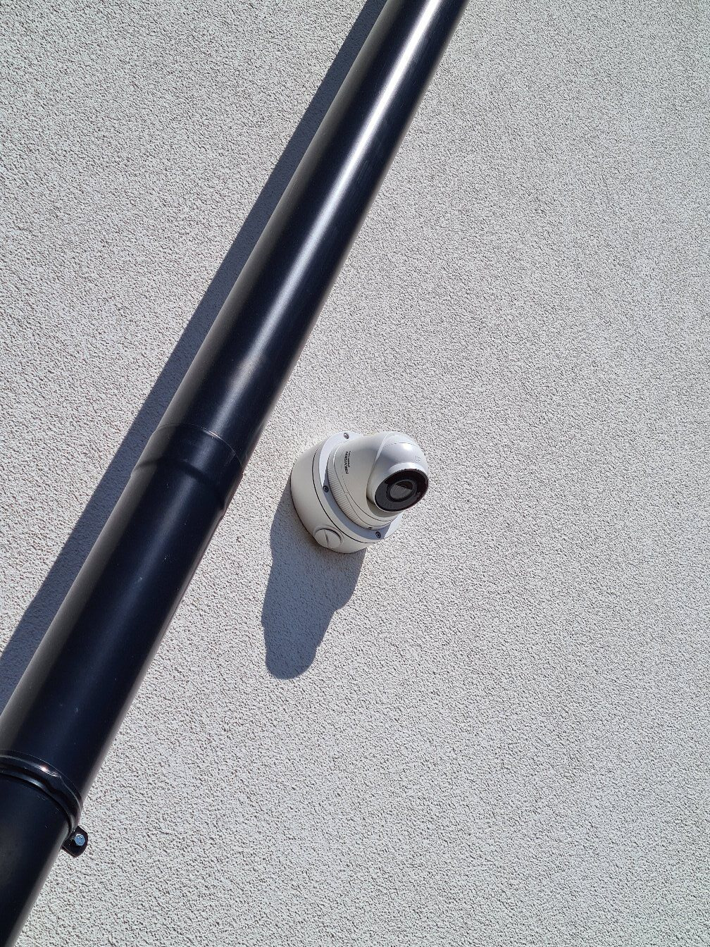 Kamera kopułkowa monitoringu Hikvision zamontowana na teksturowanej ścianie, obok czarnej rury spustowej. Jasne, naturalne oświetlenie.