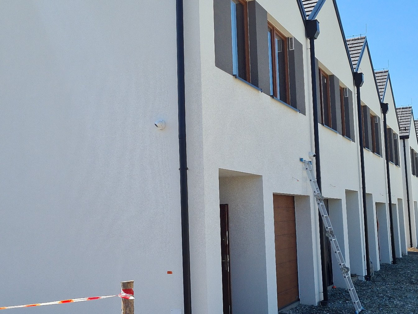 Nowoczesne segmenty mieszkalne z zamontowanym monitoringiem, widok z zewnątrz. Biała elewacja, grafitowe rynny i okna. Kamera na ścianie.