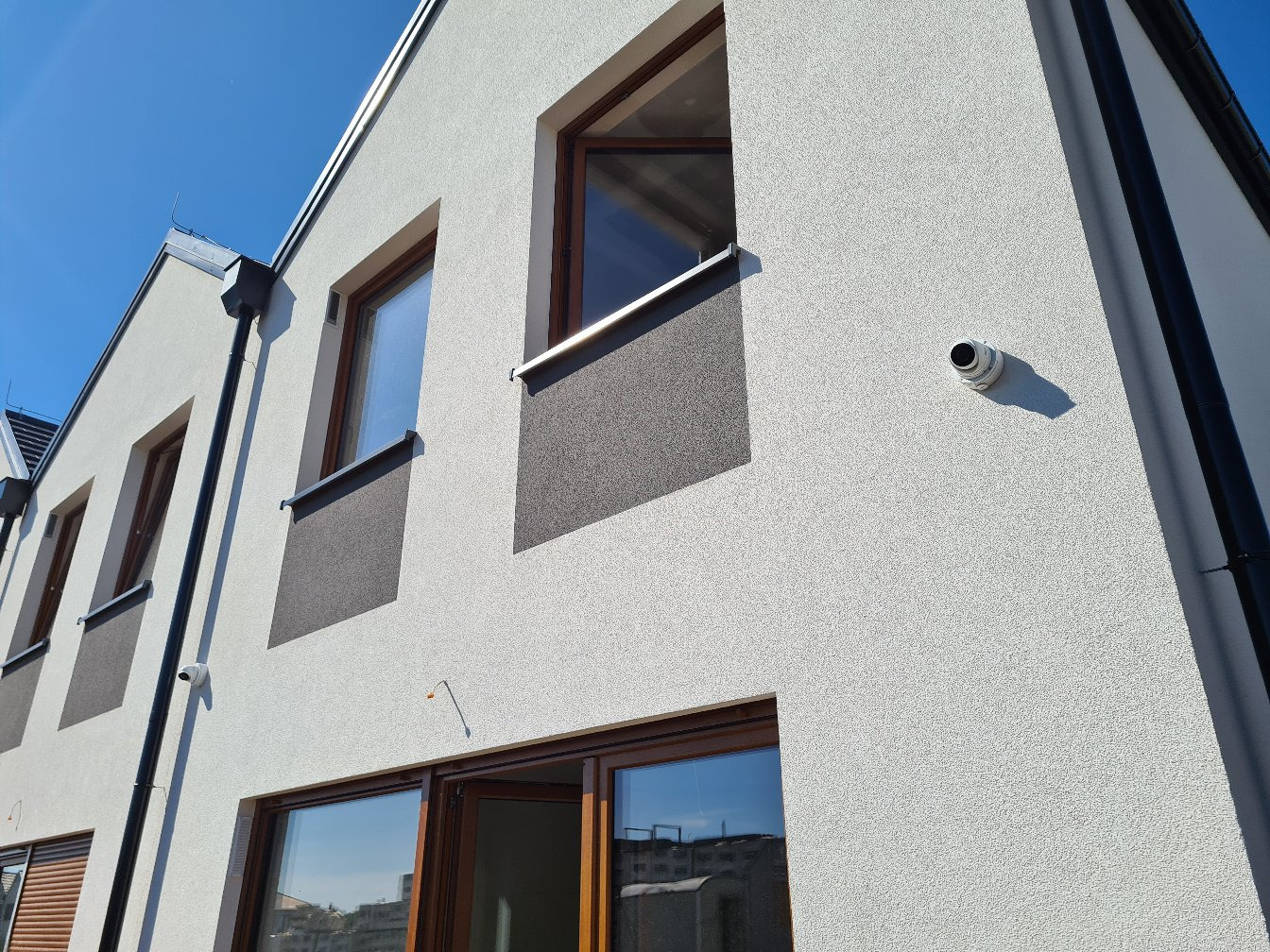 Elewacja budynku z oknami w drewnianych ramach, fragmentem dachu i zamontowaną kamerą monitoringu na białej fasadzie. Widok z dołu, błękitne niebo.