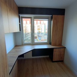 EdiMeb Meble na wymiar - Drewniane biurko na wymiar wkomponowane w przestrzeń okienną, z szafkami wiszącymi i szufladami, tworzące funkcjonalne miejsce do pracy w pokoju.