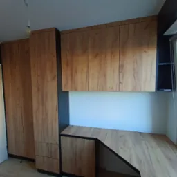 Zabudowa wnęki z biurkiem i szafkami wiszącymi, wykończona okleiną drewnopodobną w ciepłym odcieniu, z czarnymi akcentami. Widoczne drzwi z przeszkleniami i okno.