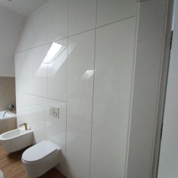 EdiMeb Meble na wymiar - Biała, połyskująca szafa wnękowa w łazience na poddaszu ze skosem, z widocznym oknem dachowym i sanitariatami. Minimalistyczny design.