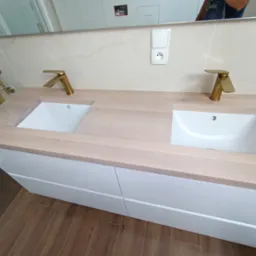 Nowoczesna łazienka z drewnianym blatem, dwiema białymi umywalkami i złotymi bateriami. Białe szafki pod blatem, płytki imitujące marmur na ścianie i drewnopodobna podłoga.