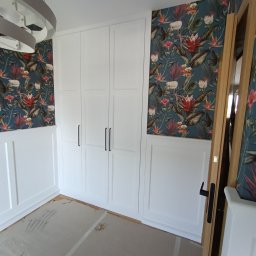 EdiMeb Meble na wymiar - Biała szafa wnękowa z czarnymi uchwytami na tle ściany z dekoracyjną tapetą w kwiaty i panele ścienne. Widoczny fragment podłogi w trakcie prac wykończeniowych.