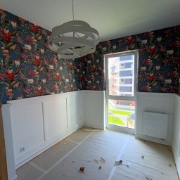 EdiMeb Meble na wymiar - Pokój po remoncie: biała boazeria, wzorzysta tapeta w tropikalne motywy, nowoczesna lampa. Widok z okna na budynek mieszkalny i zieleń. Podłoga zabezpieczona folią.