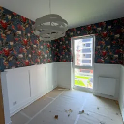 Pokój po remoncie: biała boazeria, wzorzysta tapeta w tropikalne motywy, nowoczesna lampa. Widok z okna na budynek mieszkalny i zieleń. Podłoga zabezpieczona folią.