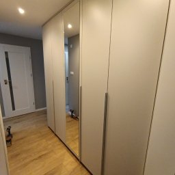 EdiMeb Meble na wymiar - Jasna szafa wnękowa z lustrzanymi drzwiami, widoczna na tle drewnianej podłogi. W lustrze odbija się wiertarka, co sugeruje niedawny montaż.