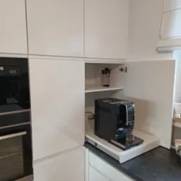 Biała, nowoczesna kuchnia z zabudowanym ekspresem do kawy na wysuwanej półce. Widoczny piekarnik i fragment blatu. Drzwiczki szafki otwarte.