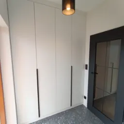 Biała szafa wnękowa z długimi uchwytami w przedpokoju, widok z dołu, w tle czarne drzwi ze szprosami. Realizacja w Rzemieniu.
