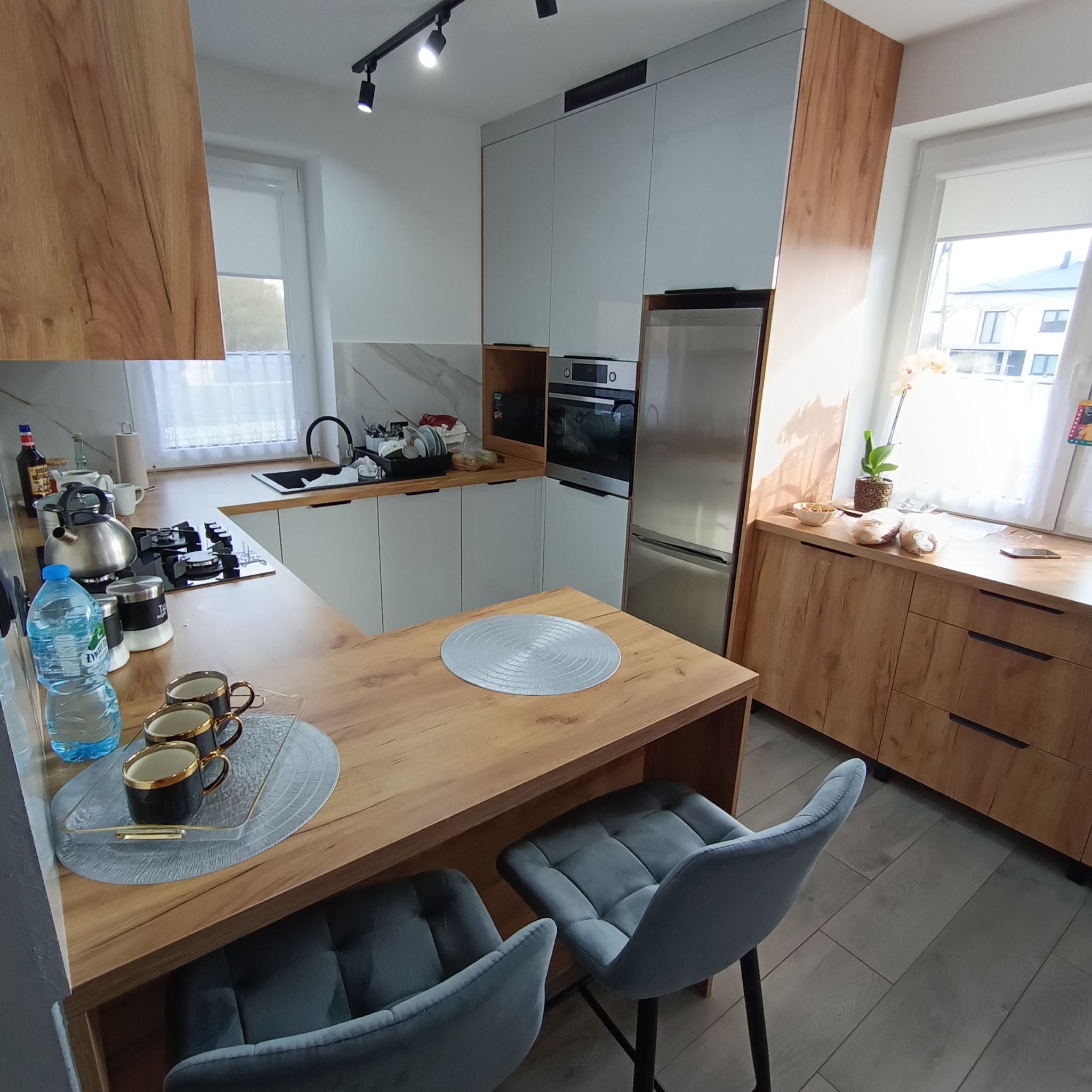 Nowoczesna kuchnia z drewnianymi akcentami: białe szafki, blat w kolorze drewna, srebrna lodówka i szare krzesła barowe. Widok na okno i elementy dekoracyjne.