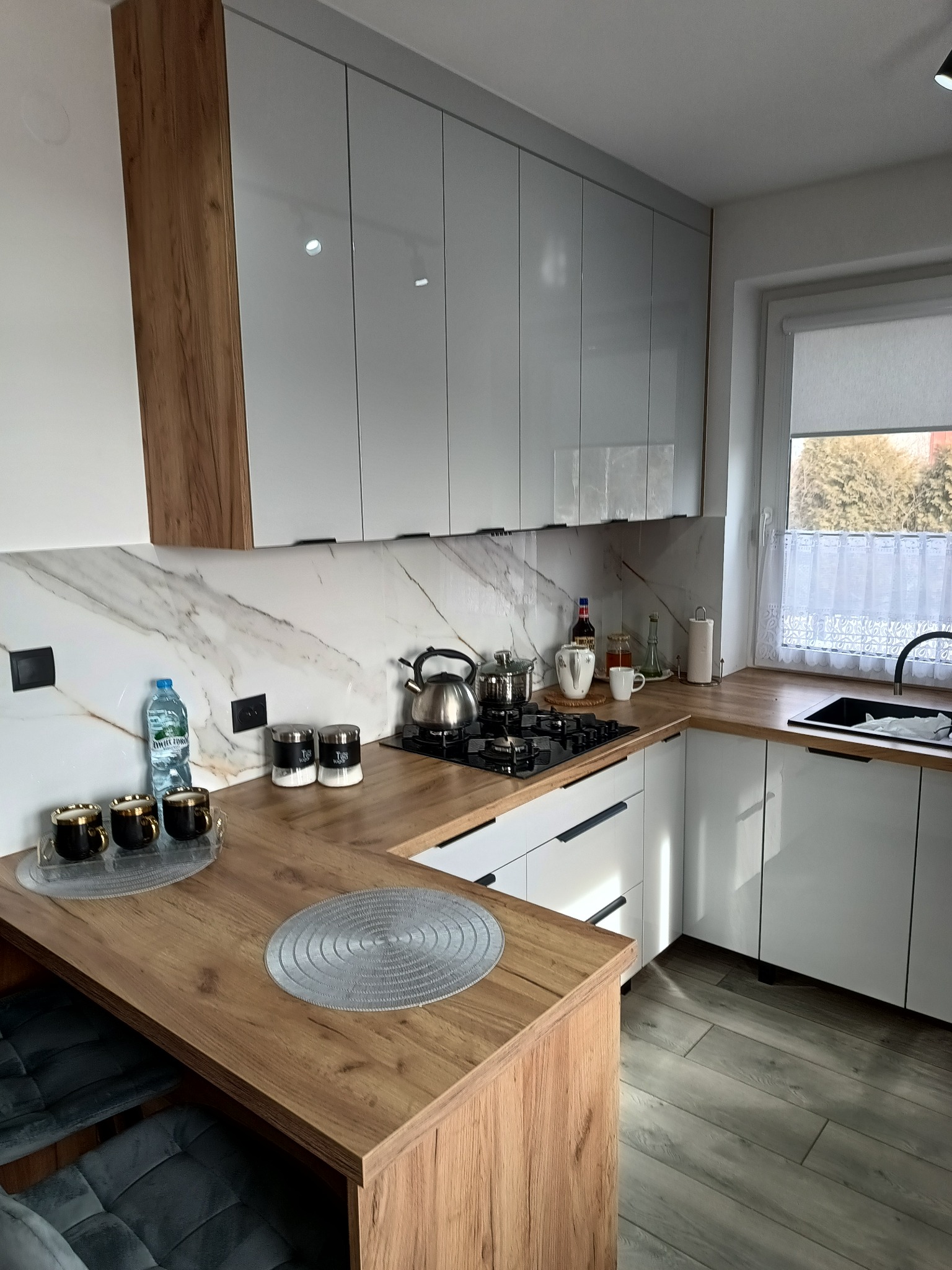 Nowoczesna kuchnia z białymi, błyszczącymi szafkami, drewnianym blatem i marmurowym backsplash'em. Na blacie czajnik, przyprawy i dekoracje. Widok na okno z firanką.