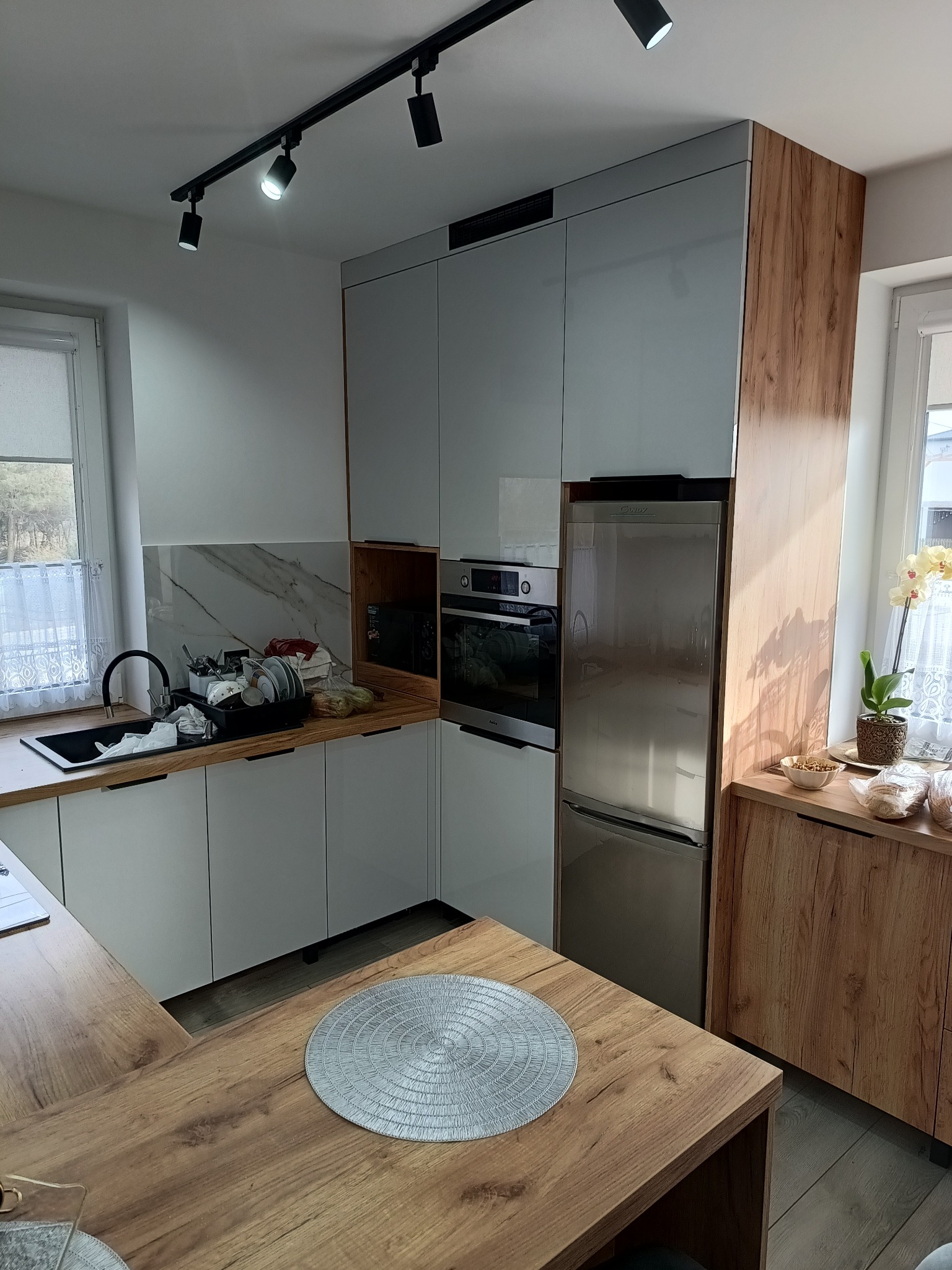 Nowoczesna kuchnia z białymi frontami i drewnianymi akcentami. Widoczny blat, szafki, sprzęt AGD oraz oświetlenie szynowe. Umywalka z naczyniami.