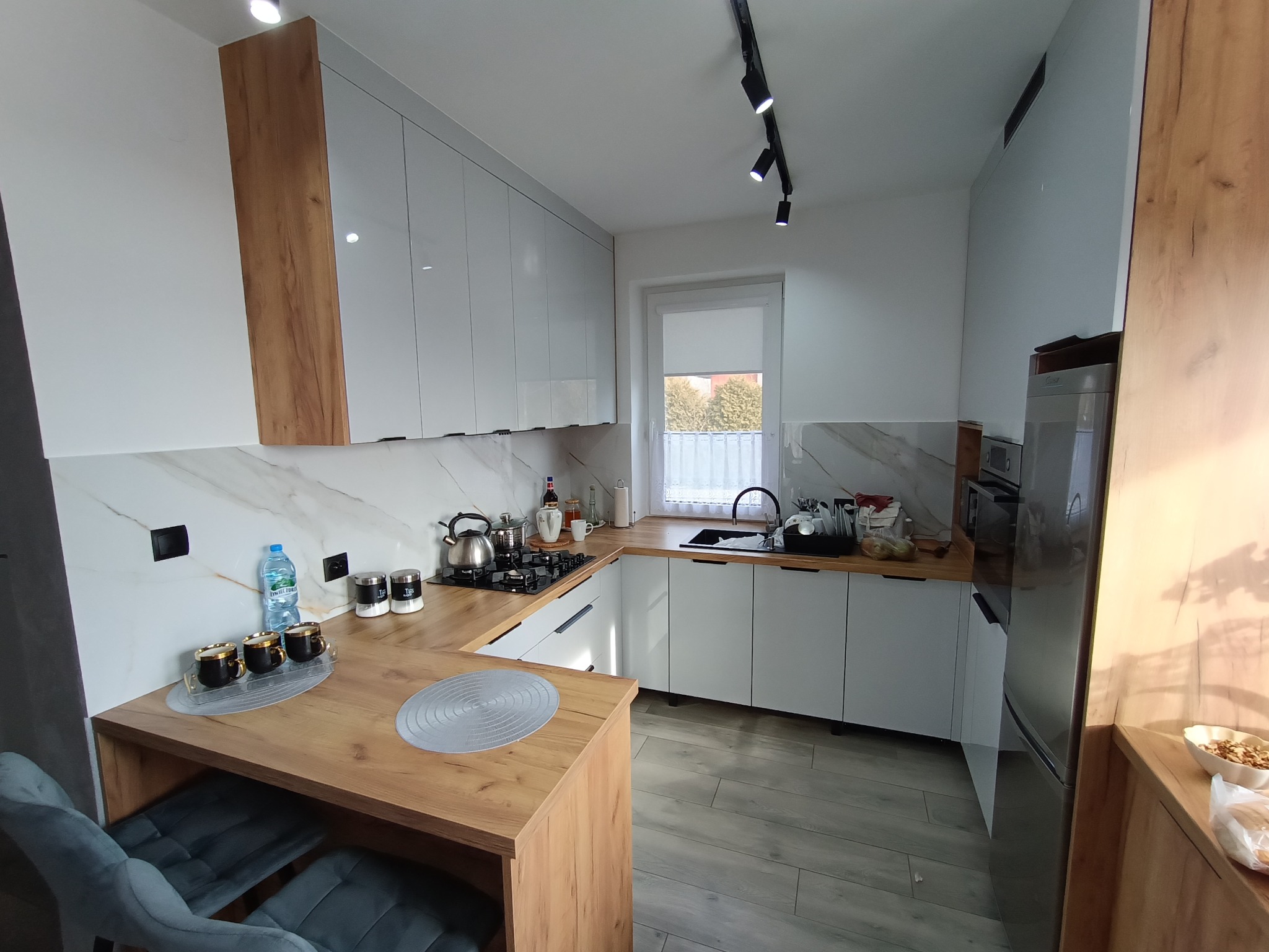 Jasna kuchnia z białymi szafkami i drewnianymi blatami, oświetlona szynoprzewodem. Widoczny stół barowy z krzesłami i elementy wyposażenia.