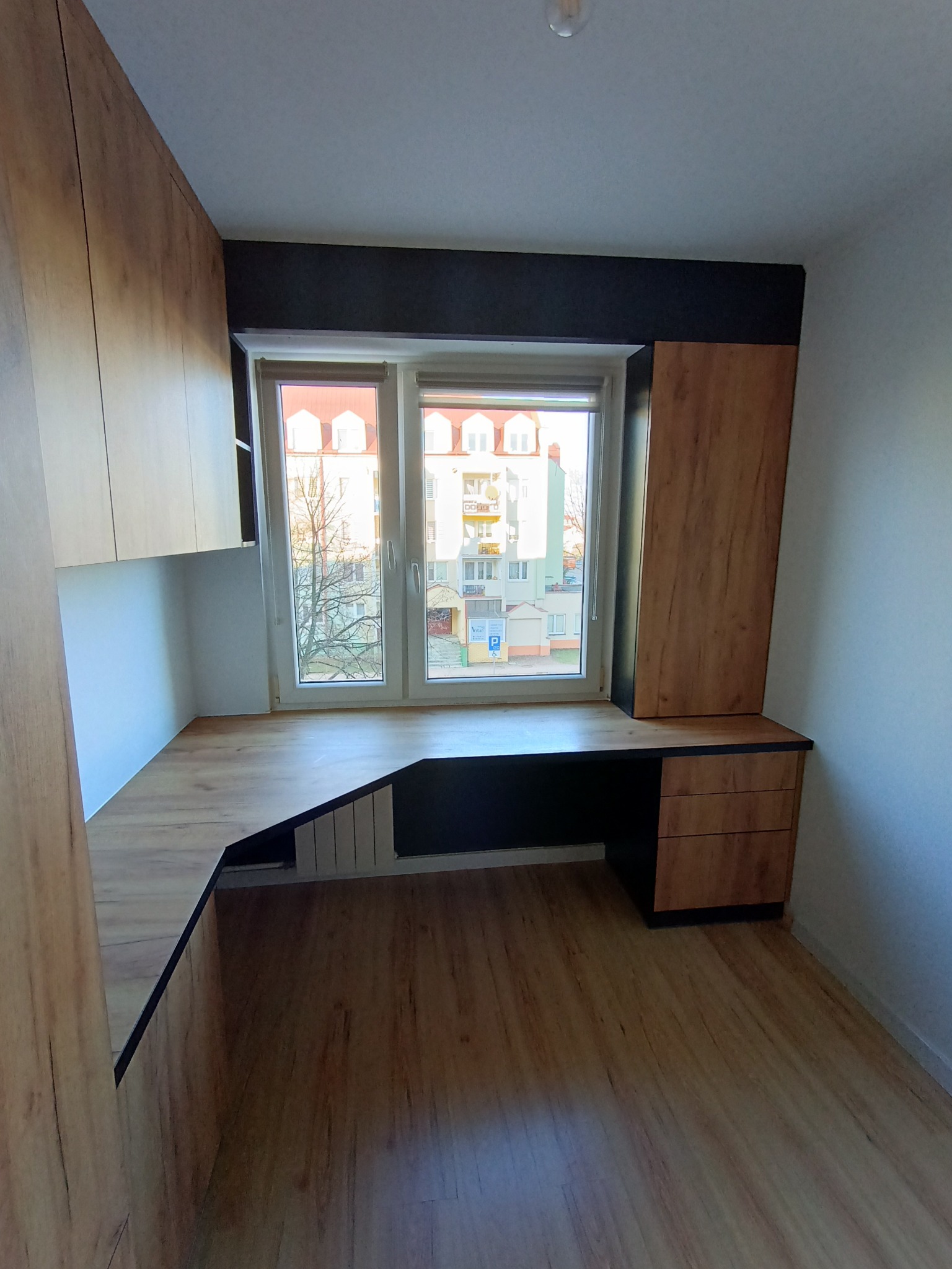 Drewniane biurko na wymiar wkomponowane w przestrzeń okienną, z szafkami wiszącymi i szufladami, tworzące funkcjonalne miejsce do pracy w pokoju.