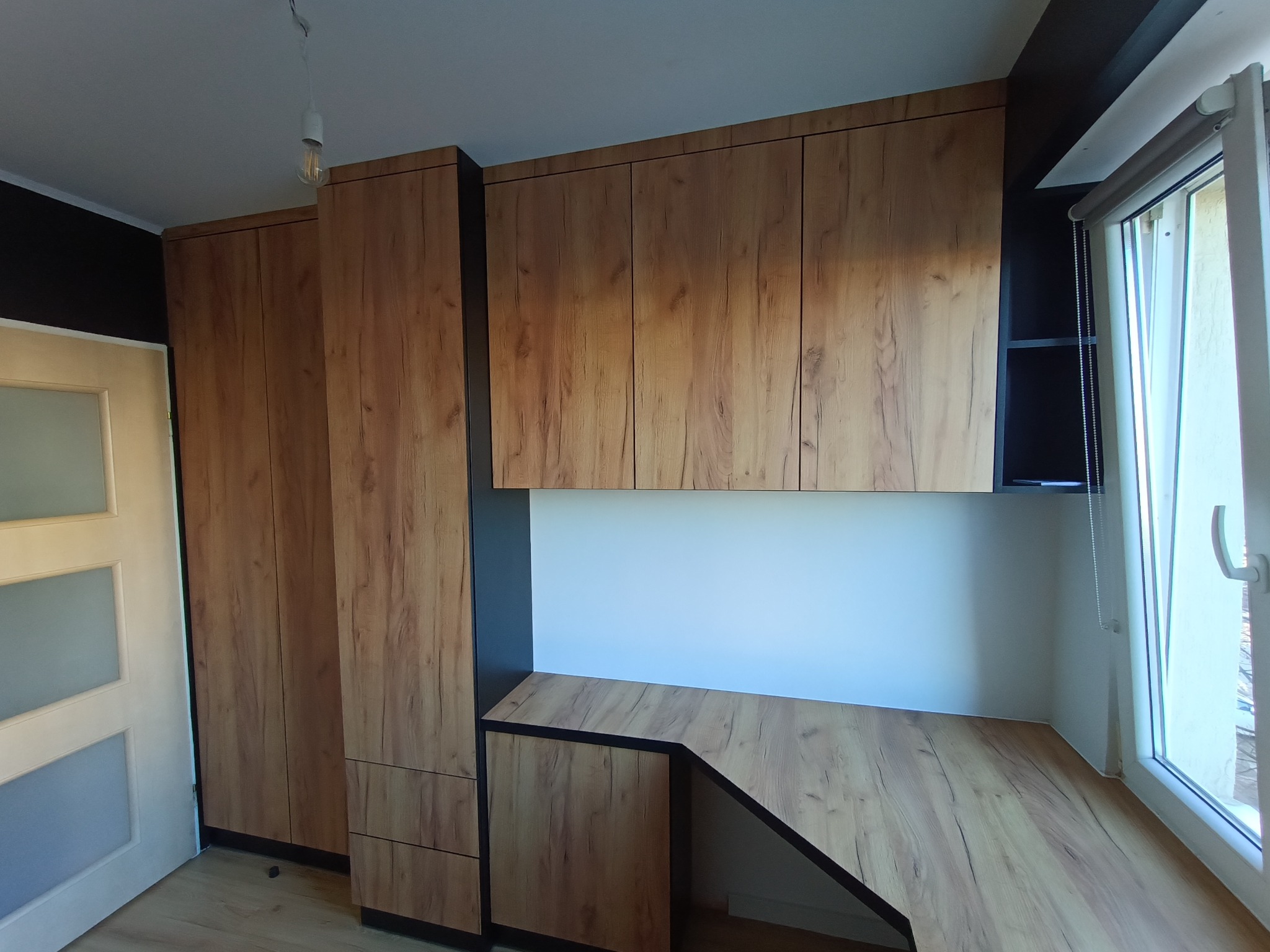 Zabudowa wnęki z biurkiem i szafkami wiszącymi, wykończona okleiną drewnopodobną w ciepłym odcieniu, z czarnymi akcentami. Widoczne drzwi z przeszkleniami i okno.