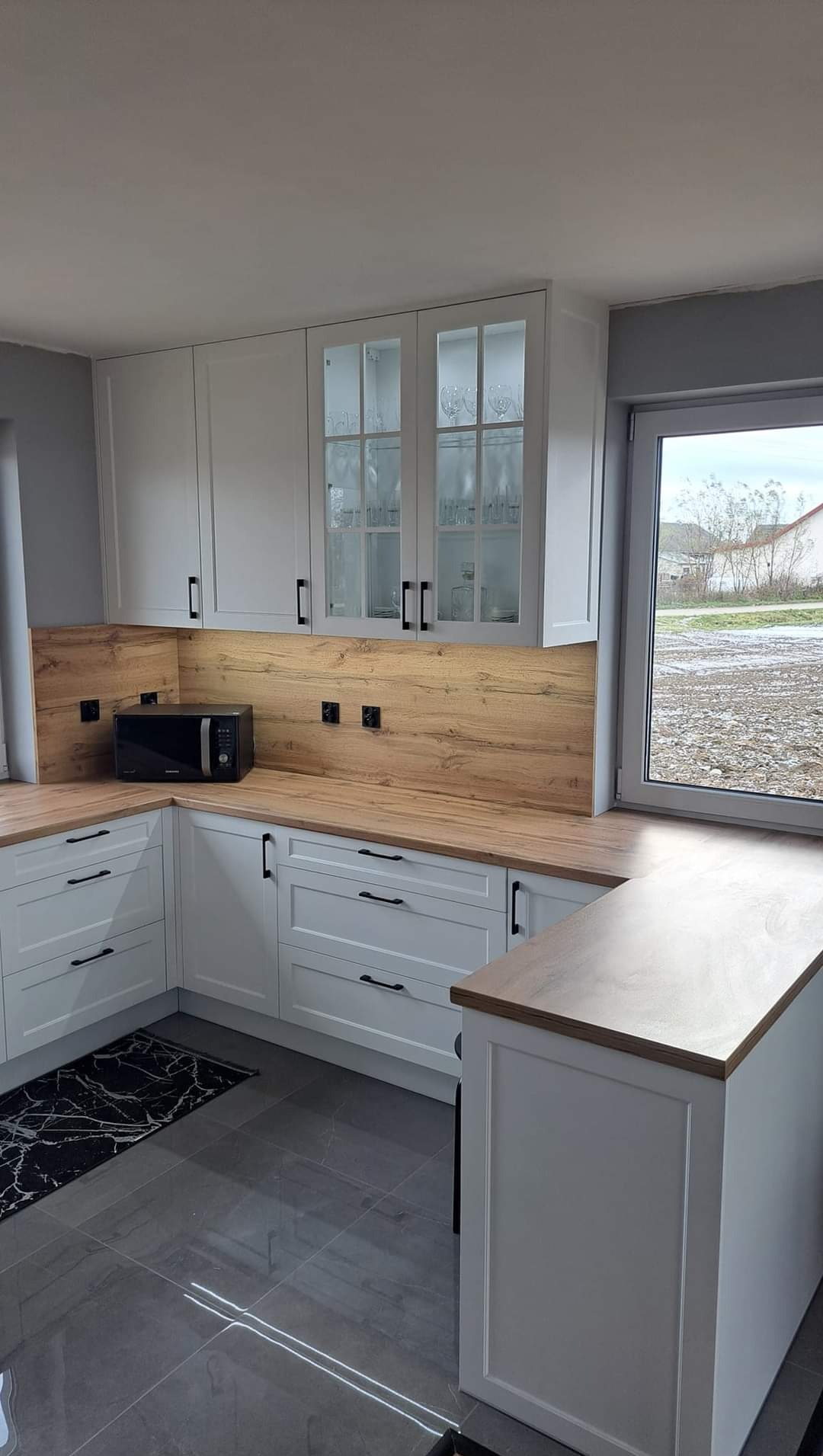 Jasna kuchnia z drewnianym blatem i backsplashiem, czarne uchwyty, szklane witryny górnych szafek, widok na zewnątrz przez okno.