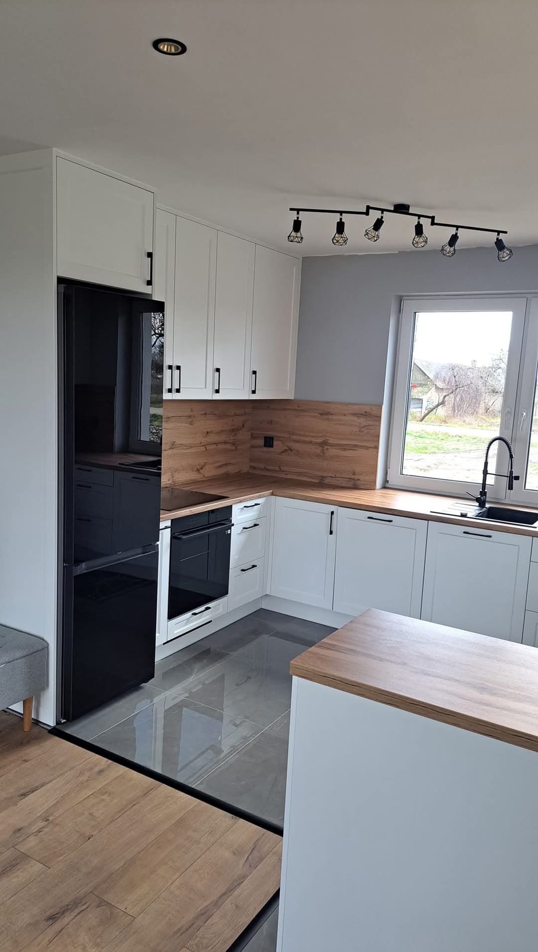 Nowoczesna, jasna kuchnia z białymi szafkami, drewnianym blatem i czarnymi akcentami, takimi jak armatura i oświetlenie szynowe. Widoczna lodówka w zabudowie.