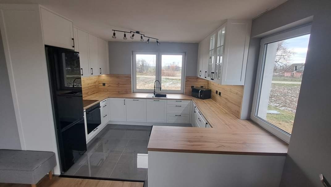 Jasna kuchnia z drewnianymi blatami i czarnym sprzętem AGD, oświetlona lampą sufitową z żarówkami. Widok z okna na zewnątrz. Nowoczesne wykończenie.