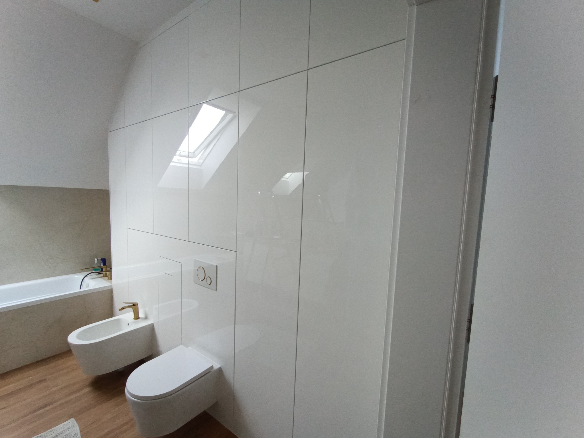 Biała, połyskująca szafa wnękowa w łazience na poddaszu ze skosem, z widocznym oknem dachowym i sanitariatami. Minimalistyczny design.