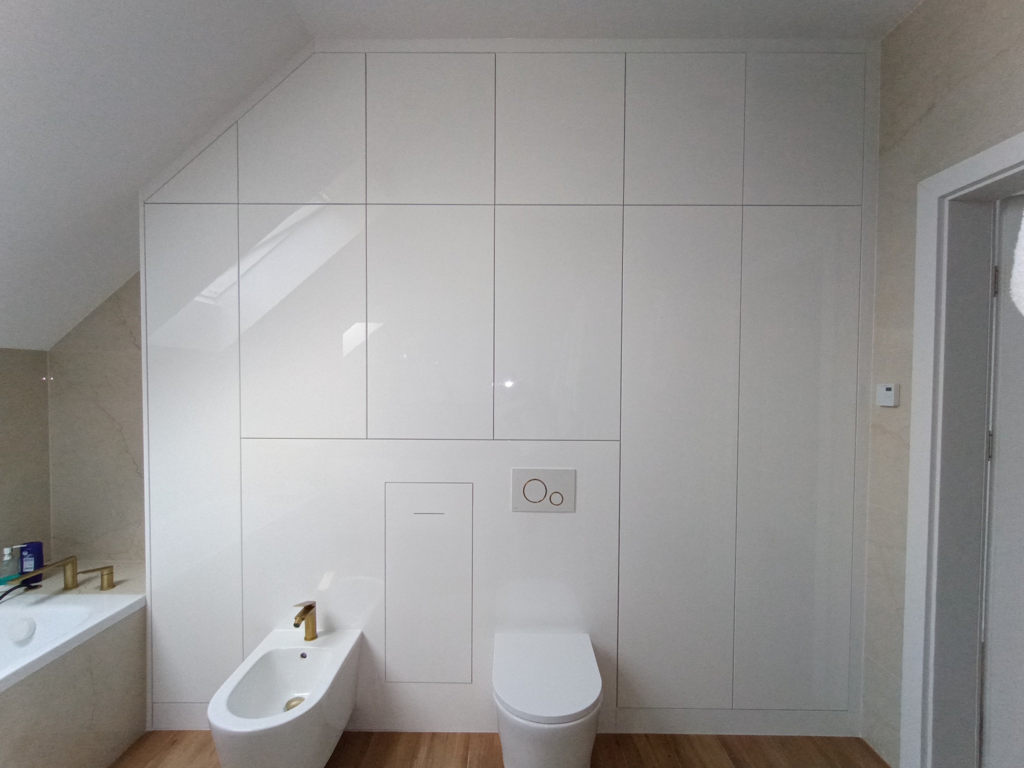 Biała, lakierowana szafa wnękowa pod skosem w łazience, z ukrytą spłuczką WC i bidetem. Złota armatura. Drewnopodobna podłoga. Minimalistyczny design.