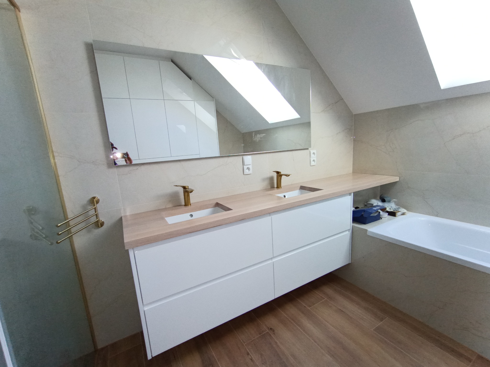 Nowoczesna łazienka na poddaszu z dwiema umywalkami nablatowymi i drewnianym blatem. W lustrze odbija się biała szafa w skosie. Złota armatura i drewnopodobna podłoga.
