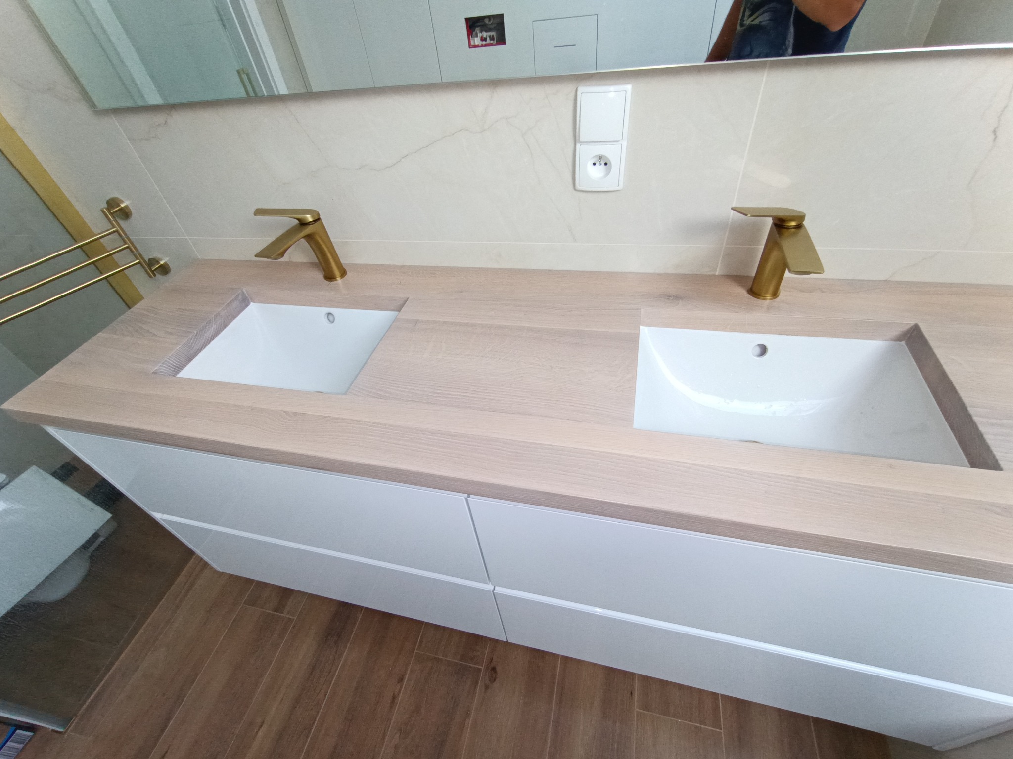 Nowoczesna łazienka z drewnianym blatem, dwiema białymi umywalkami i złotymi bateriami. Białe szafki pod blatem, płytki imitujące marmur na ścianie i drewnopodobna podłoga.