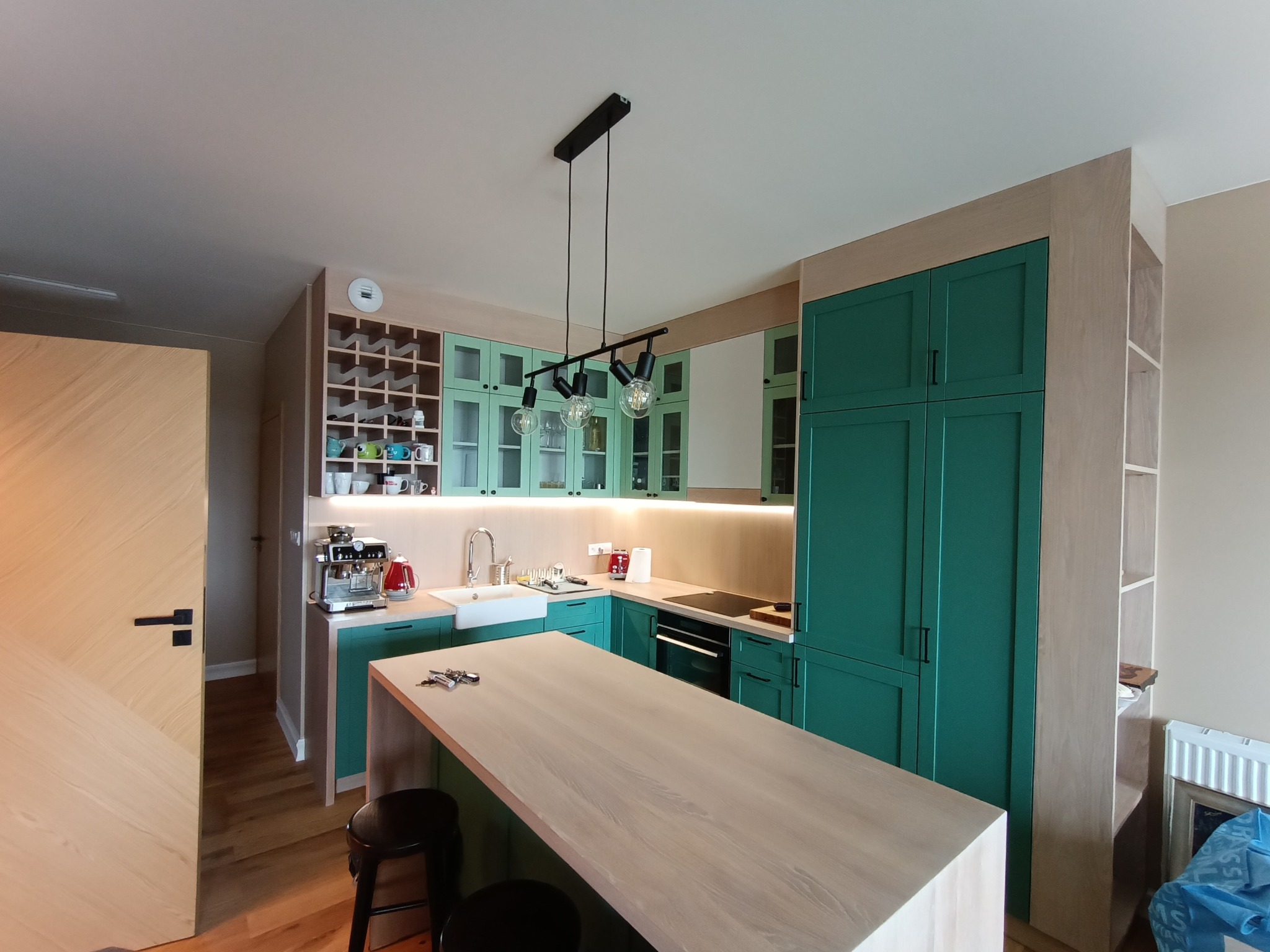 Nowoczesna kuchnia z wyspą i zielonymi szafkami, oświetlona lampą wiszącą. Widoczny ekspres do kawy i półki na wino. Całość w jasnych, naturalnych barwach.
