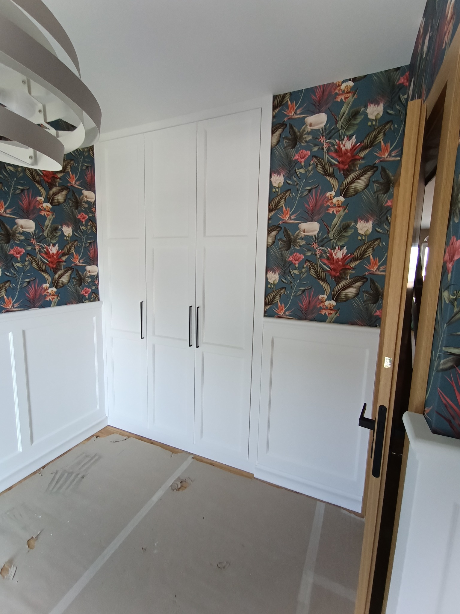 Biała szafa wnękowa z czarnymi uchwytami na tle ściany z dekoracyjną tapetą w kwiaty i panele ścienne. Widoczny fragment podłogi w trakcie prac wykończeniowych.