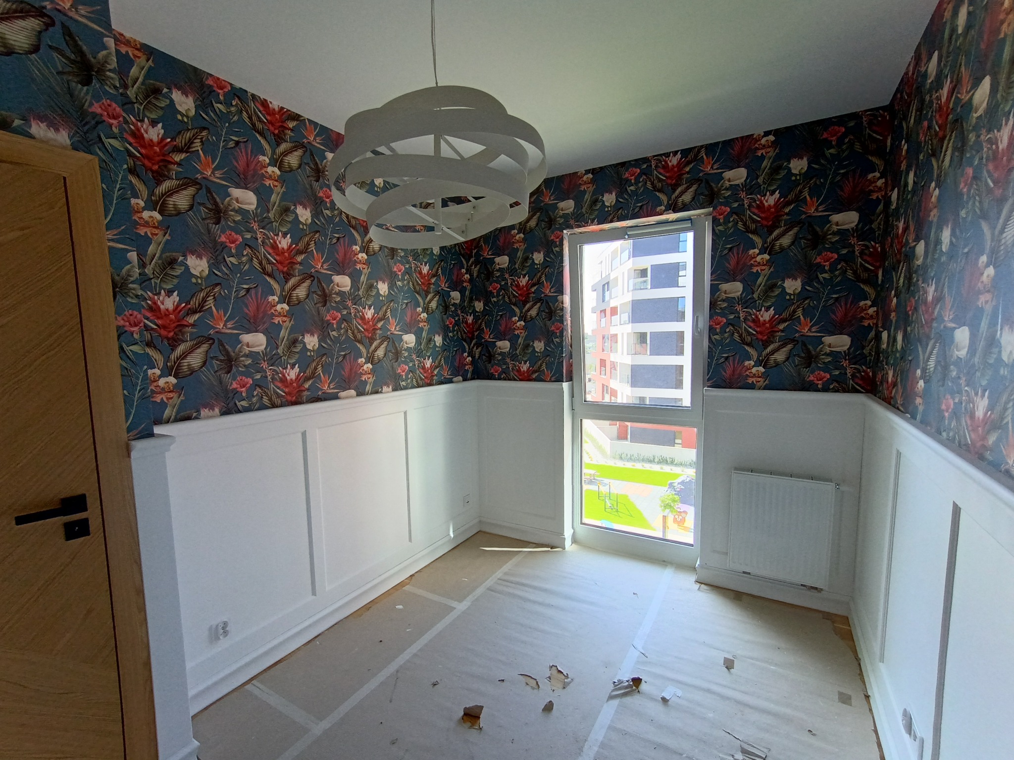 Pokój po remoncie: biała boazeria, wzorzysta tapeta w tropikalne motywy, nowoczesna lampa. Widok z okna na budynek mieszkalny i zieleń. Podłoga zabezpieczona folią.