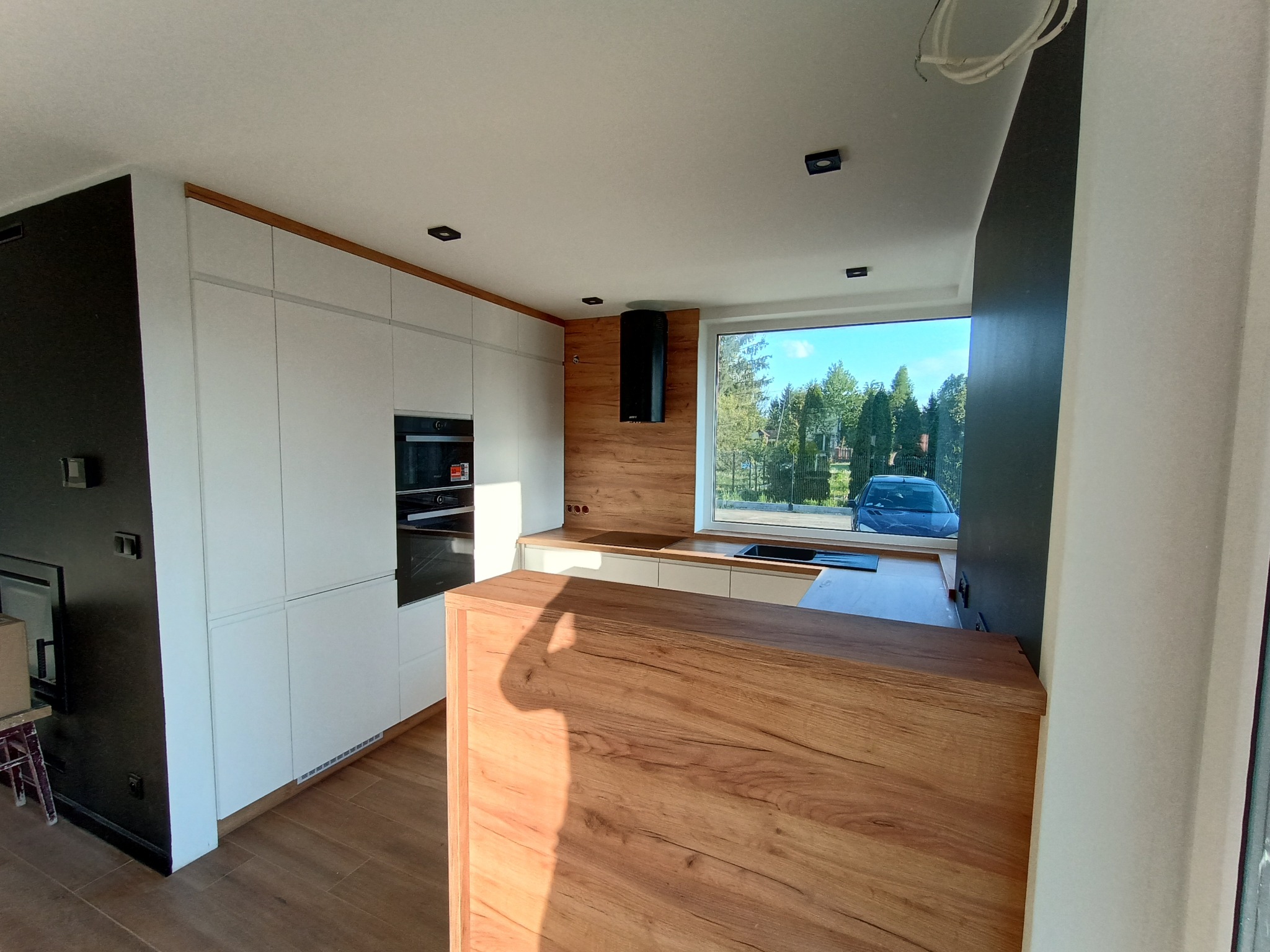 Nowoczesna kuchnia z białymi szafkami, drewnianym blatem i okapem. Widok zza drewnianej wyspy. Za oknem zieleń drzew i niebieski samochód.