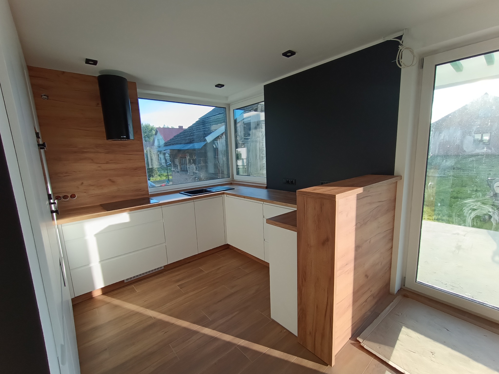 Nowoczesna kuchnia z białymi szafkami i drewnianym blatem, oświetlona światłem dziennym wpadającym przez okno z widokiem na domy. Czarny okap nad płytą.