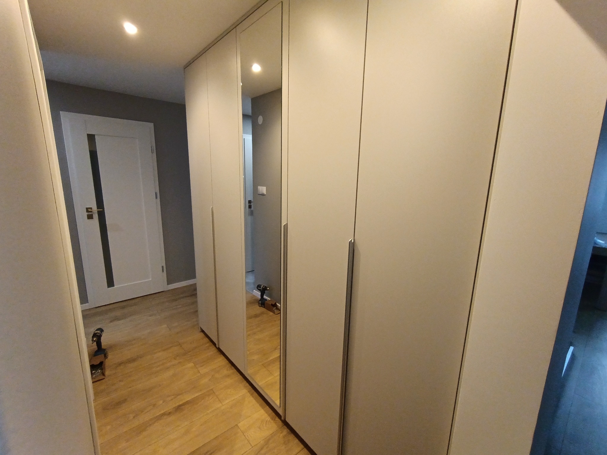 Jasna szafa wnękowa z lustrzanymi drzwiami, widoczna na tle drewnianej podłogi. W lustrze odbija się wiertarka, co sugeruje niedawny montaż.