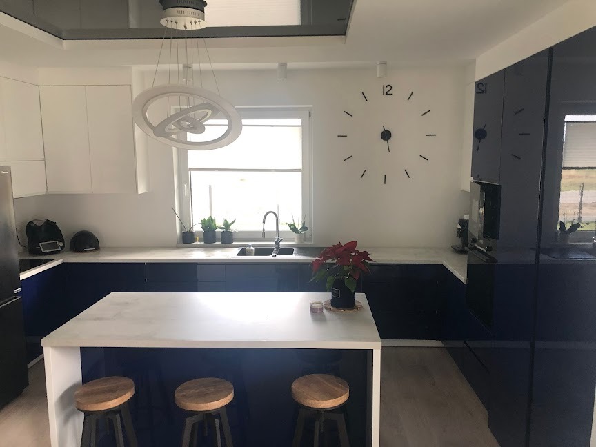 Nowoczesna kuchnia z niebieskimi szafkami na wysoki połysk, białym blatem i wyspą z drewnianymi stołkami. Na ścianie duży zegar, nad wyspą designerska lampa.