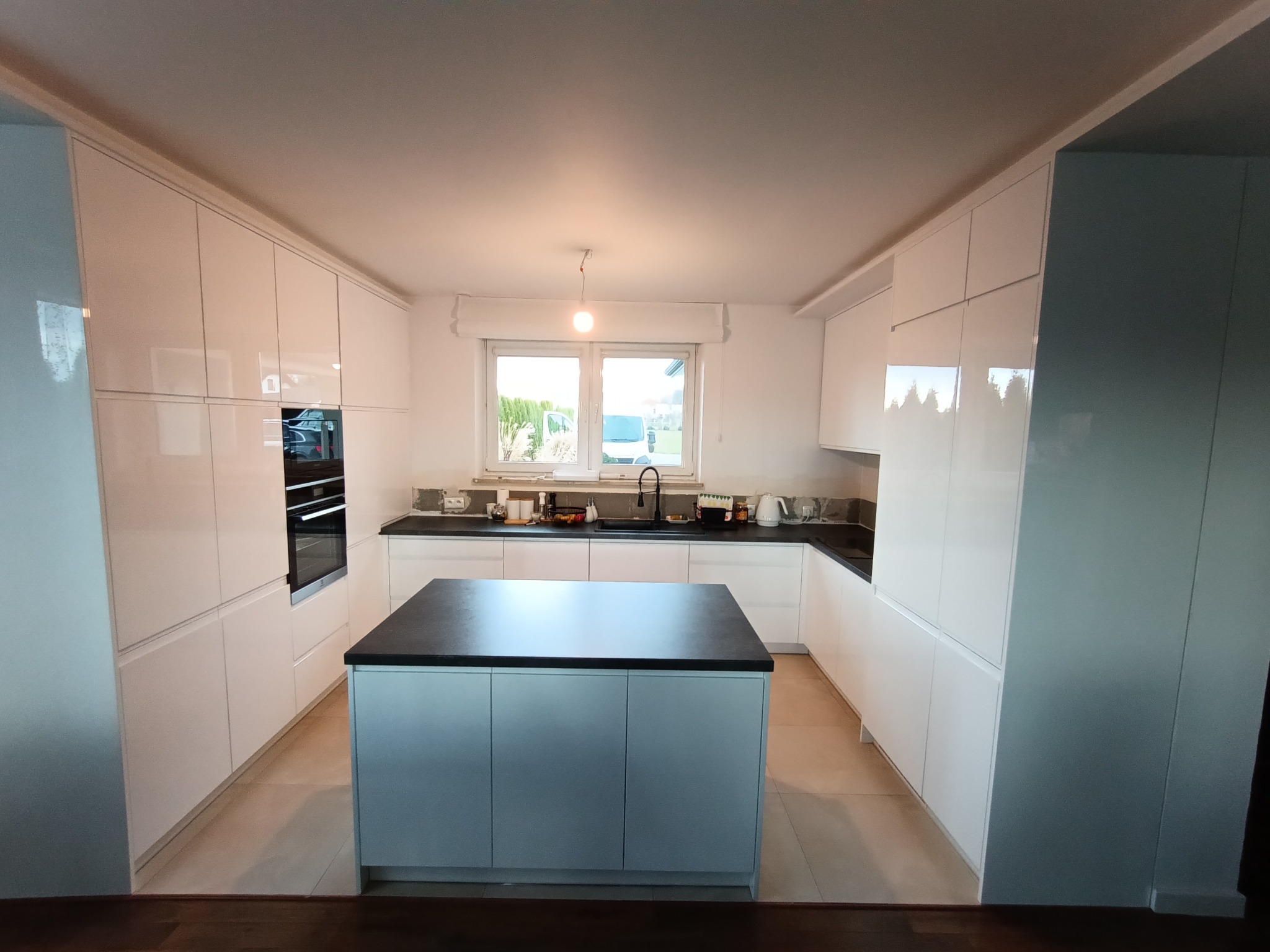 Nowoczesna, biała kuchnia z wyspą na środku i czarnym blatem. Minimalistyczny design, połyskliwe fronty szafek. Duże okno w tle, wpuszczające naturalne światło.
