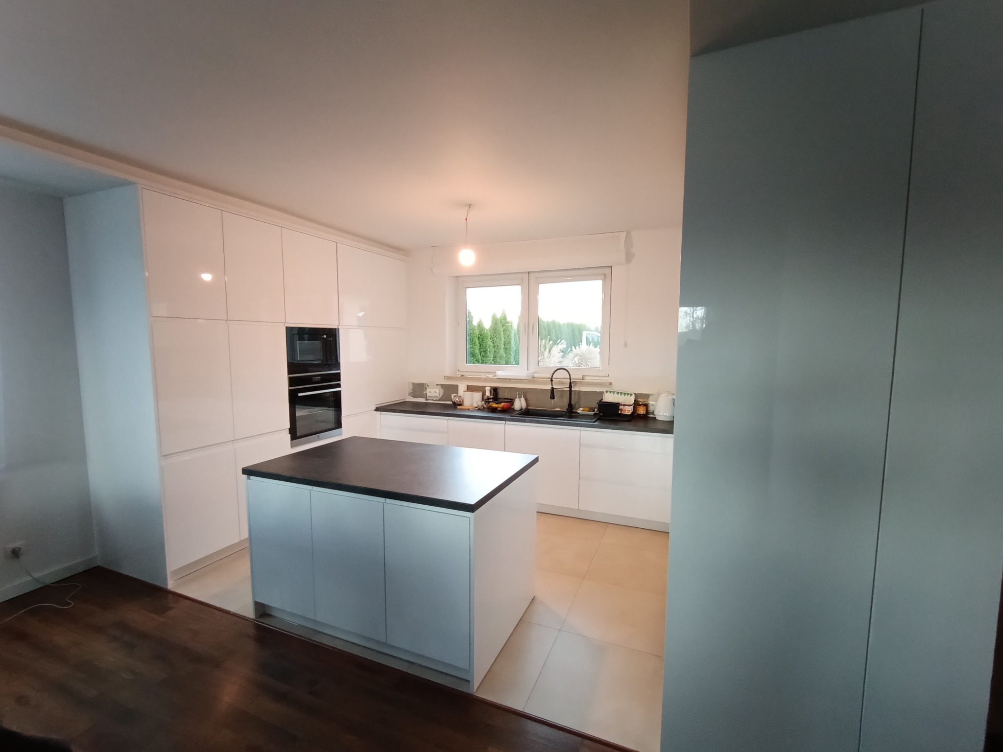 Biała kuchnia z wyspą i czarnym blatem. Nowoczesne szafki bez uchwytów, okno z widokiem na zieleń. Minimalistyczny design, jasne wnętrze, płytki podłogowe w odcieniach beżu.