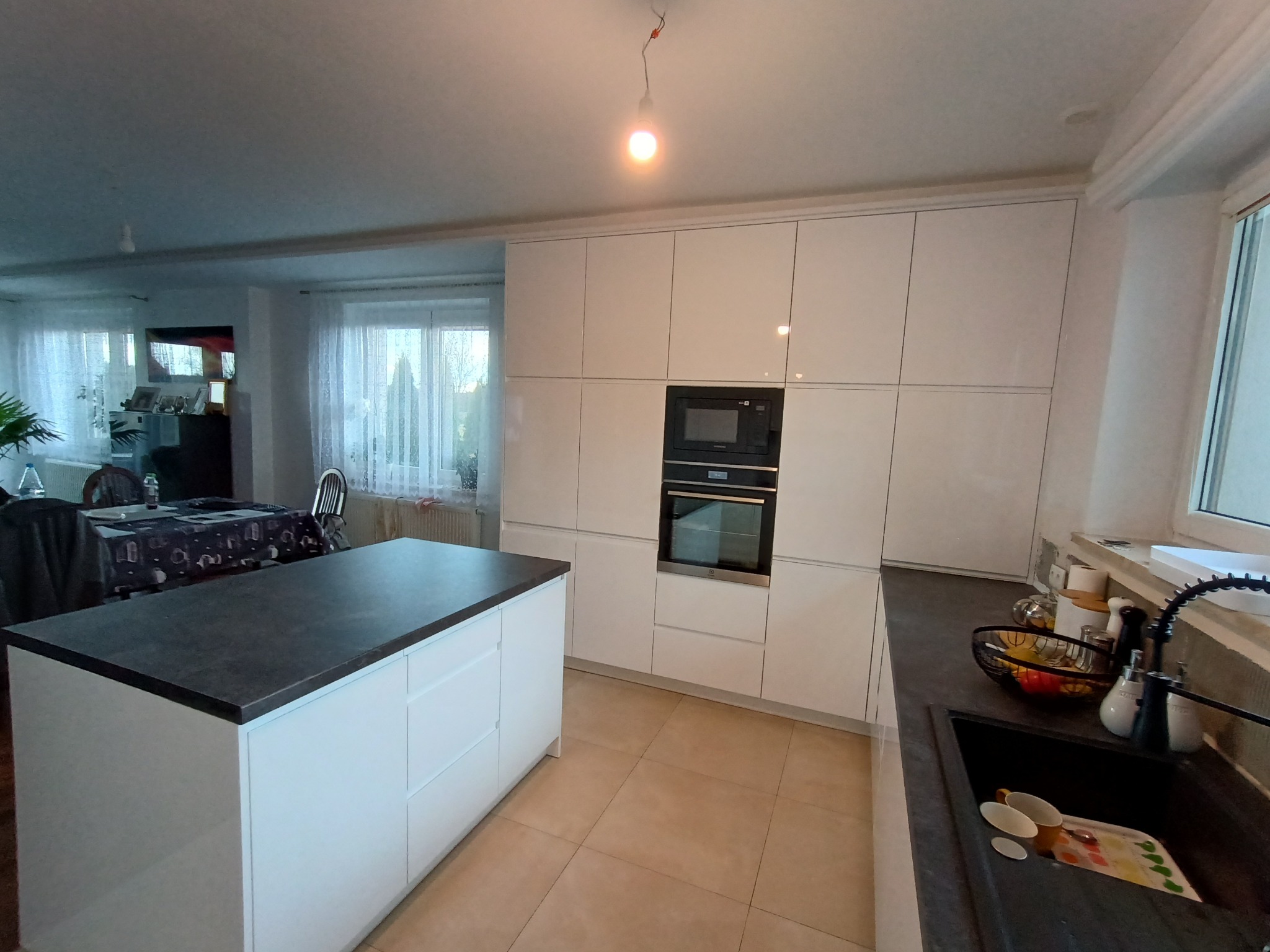 Nowoczesna, jasna kuchnia z wyspą i zabudową pod sufit. Minimalistyczne fronty szafek, ciemny blat, płytki podłogowe. Widok na stół i okno.