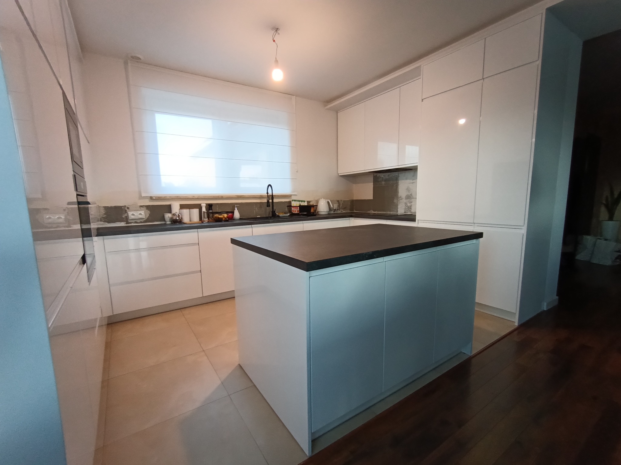 Nowoczesna, biała kuchnia z wyspą i czarnym blatem. Proste fronty szafek, minimalistyczny design. Widoczne okno z roletą rzymską i elementy wyposażenia kuchennego.