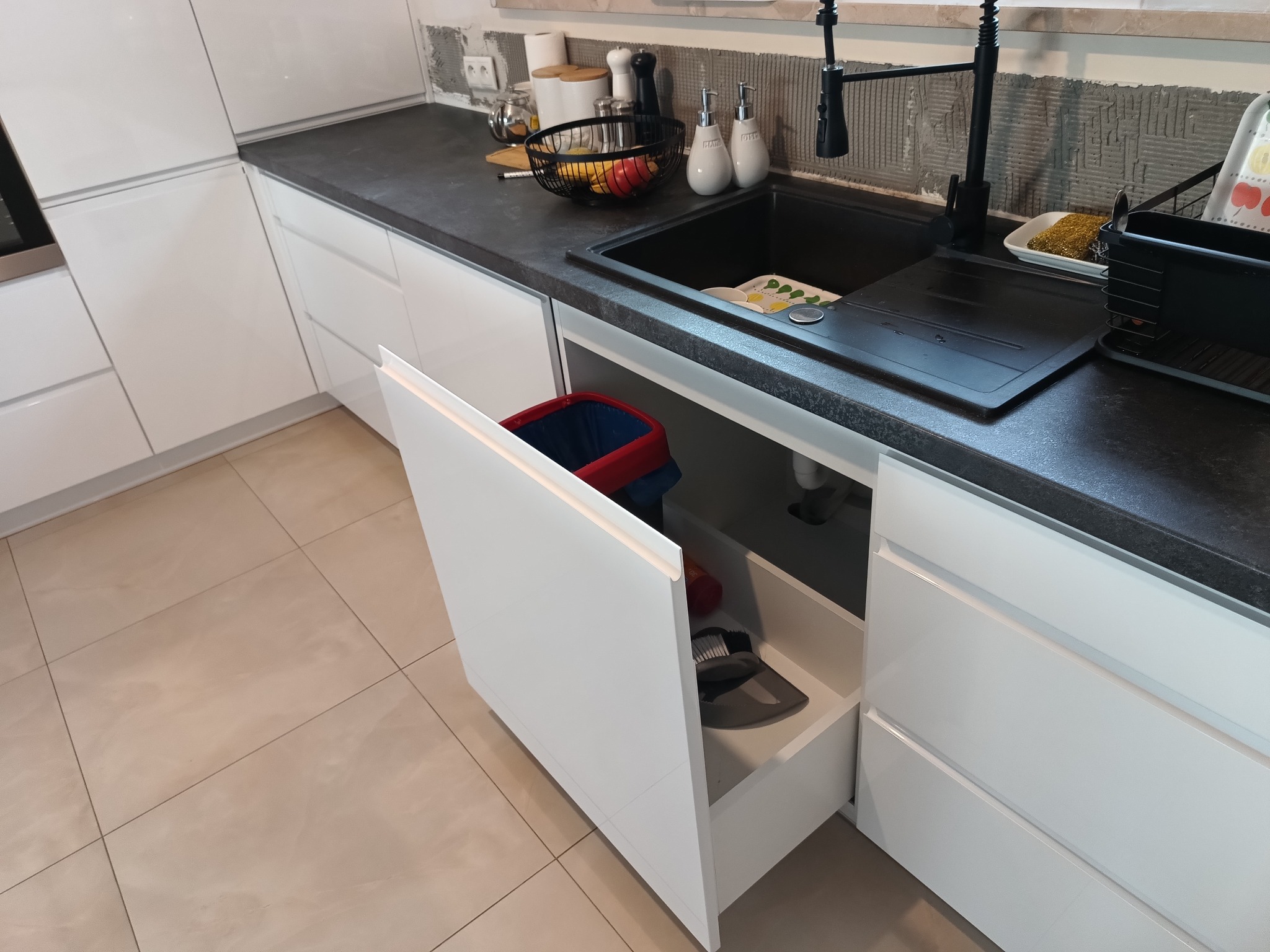 Nowoczesna, biała kuchnia z czarnym blatem i otwartą szafką pod zlewem, ukazującą kosz i akcesoria do sprzątania. Widoczne płytki na ścianie w trakcie montażu.