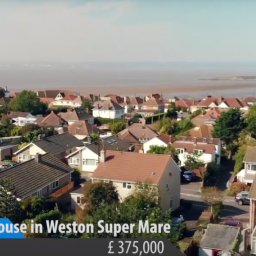 Urban Prism Media - Panoramiczny widok z drona na osiedle domów jednorodzinnych w Weston Super Mare, z widoczną linią brzegową w tle i informacją o cenie nieruchomości.