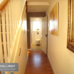 Urban Prism Media - Korytarz z drewnianymi schodami, lustrem w złotej ramie i widokiem na łazienkę. Jasne ściany, drewniana podłoga. Ujęcie z perspektywy osoby wchodzącej.