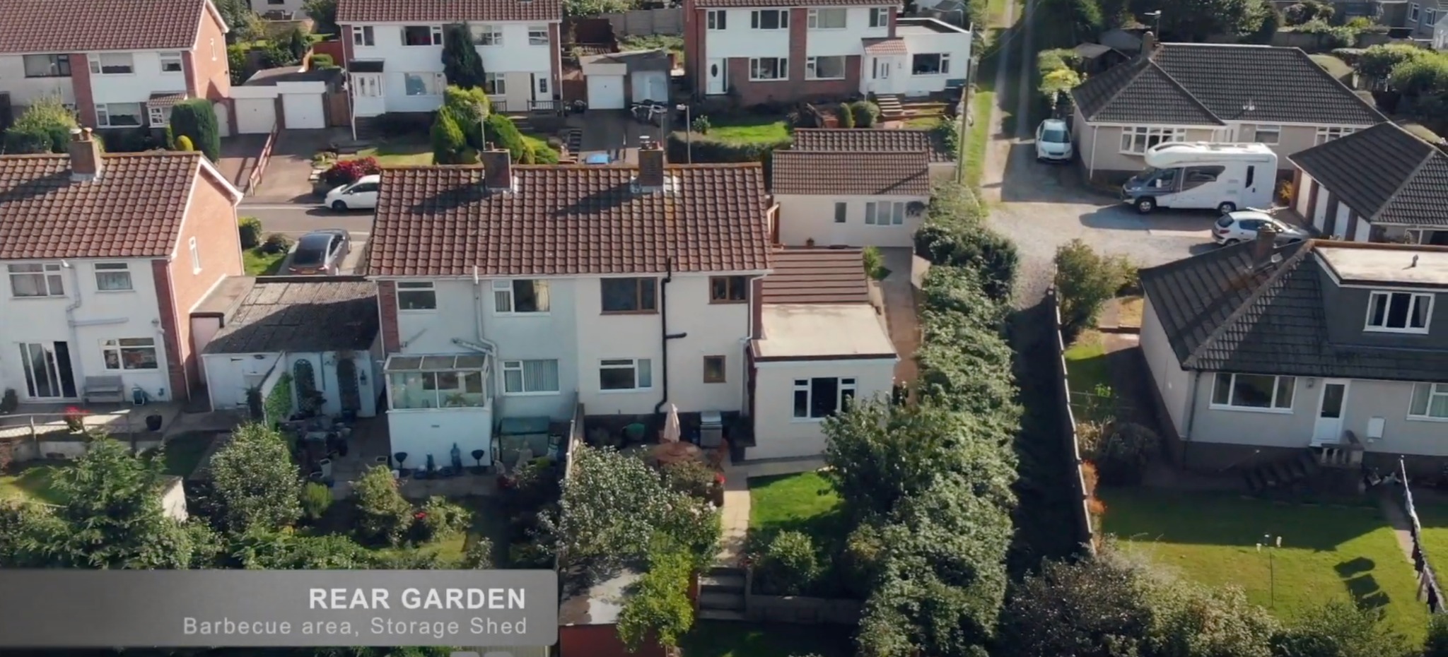 Zdjęcie z drona przedstawia osiedle domów jednorodzinnych z zadbanymi ogrodami. Widoczny kamper i samochody na podjazdach. Tło: zielone drzewa i krzewy.