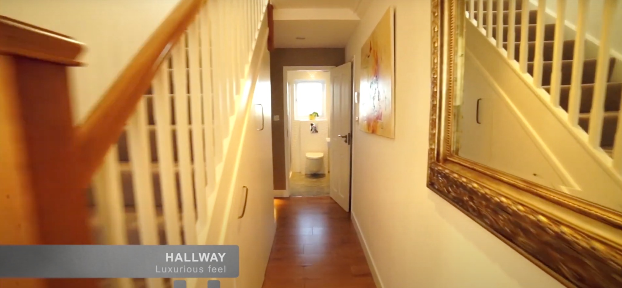 Korytarz z drewnianymi schodami, lustrem w złotej ramie i widokiem na łazienkę. Jasne ściany, drewniana podłoga. Ujęcie z perspektywy osoby wchodzącej.