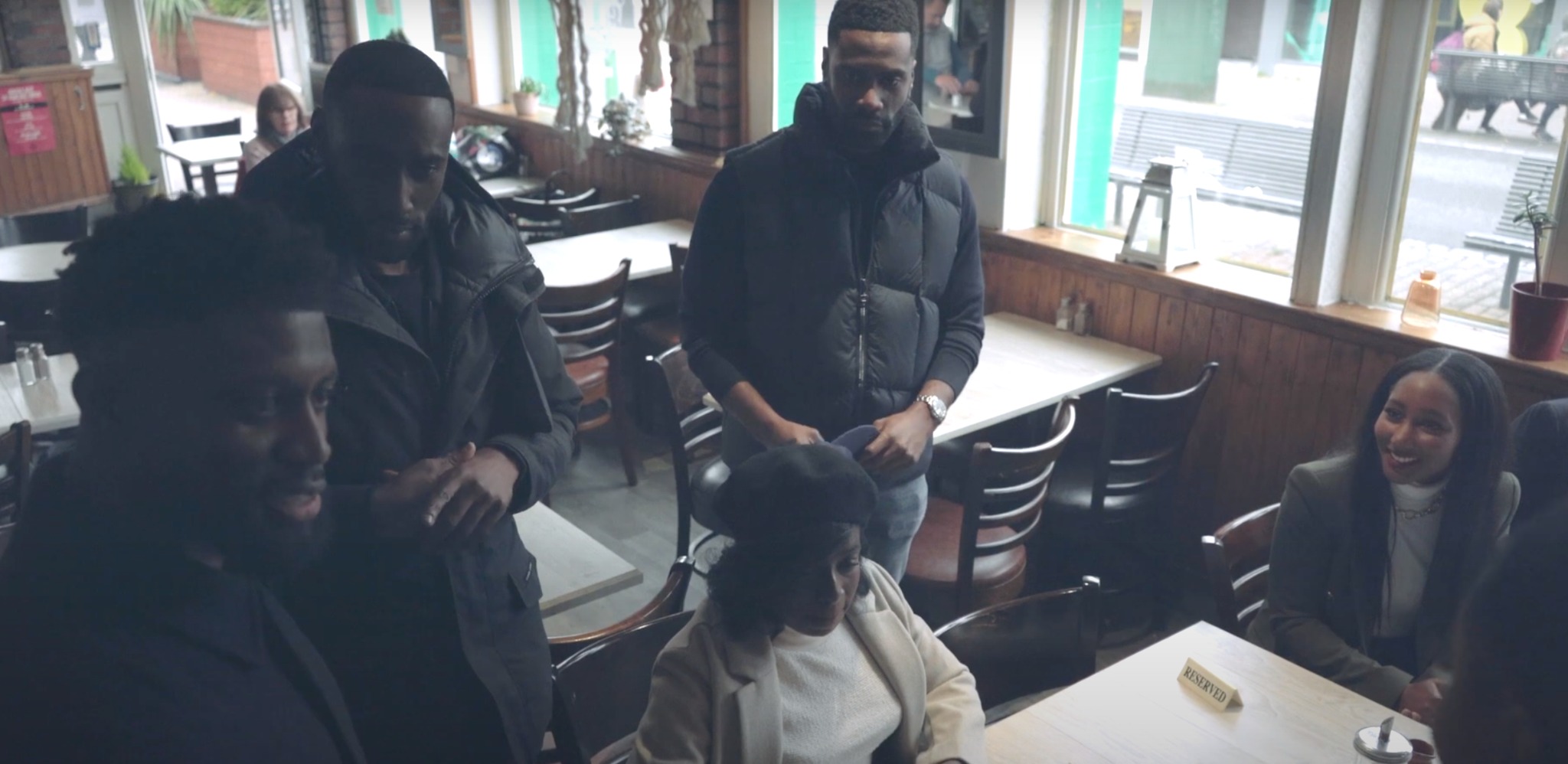 Uczestnicy eventu w kawiarni, z widoczną tabliczką 'Reserved' na stole. Mężczyźni w kurtkach i kobieta w marynarce, uśmiechnięta, siedzą przy stolikach.